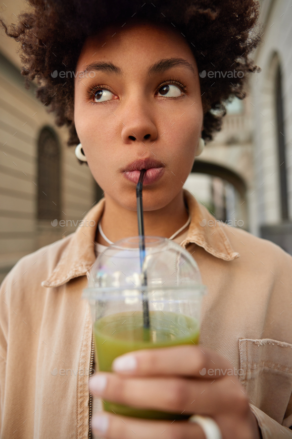 Close up shot of pensive curly teenage girl drinks green juice